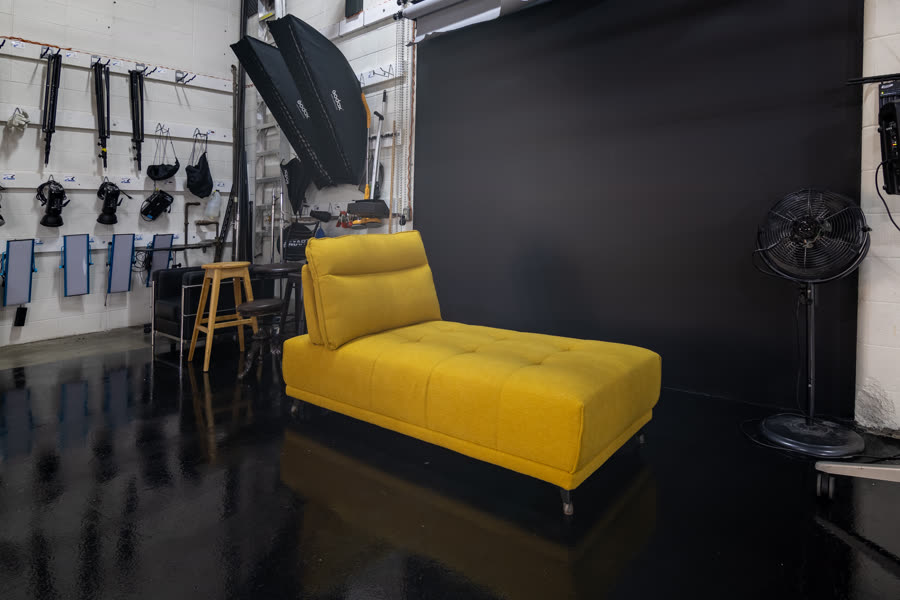 Studio A's black wall set, a mustard chaise lounge in front of a black backdrop with grip and lighting gear along the side wall
