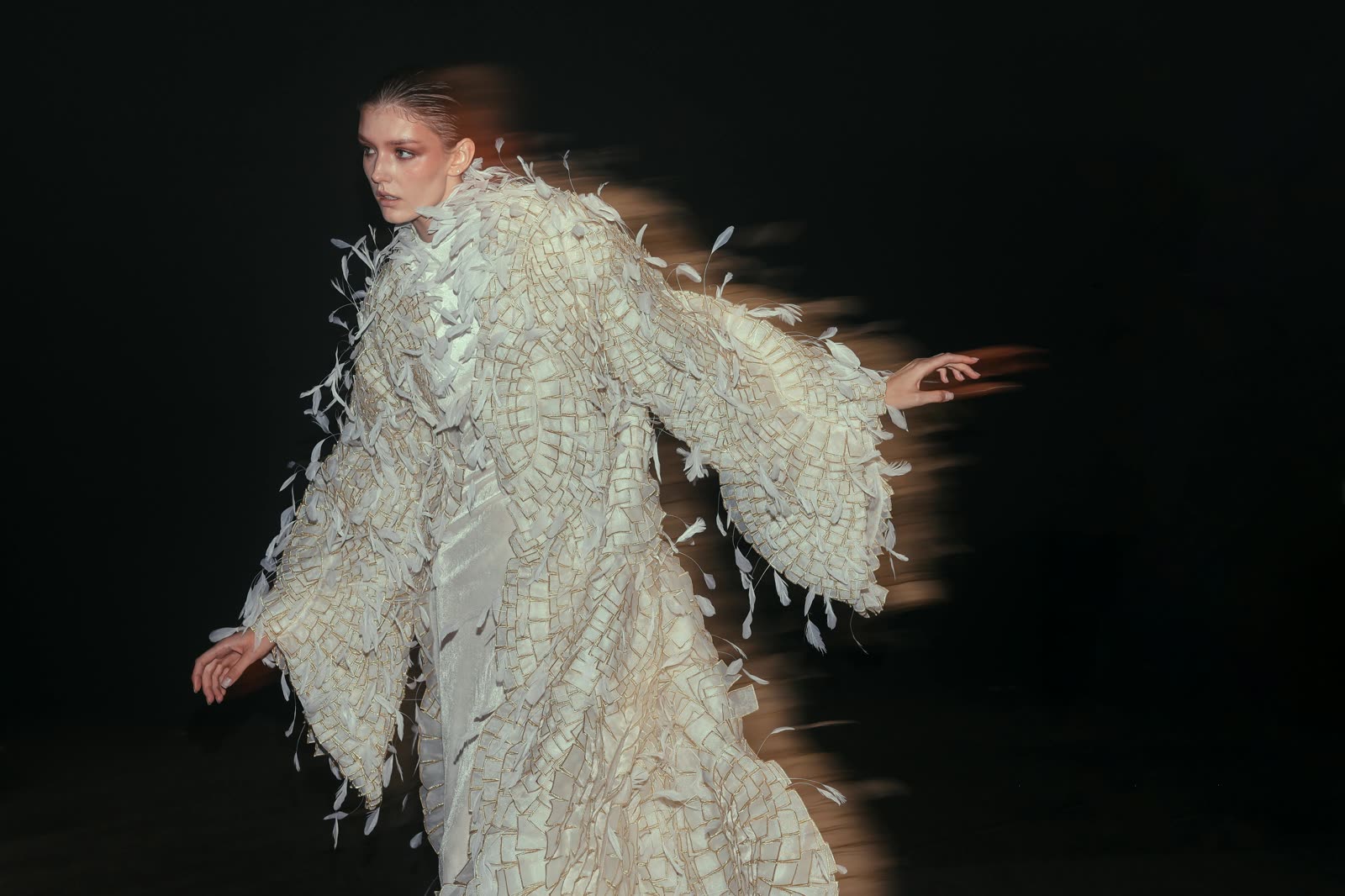 A model in a feather and gold embellished gown captured mid-movement against a dark backdrop at Studio A