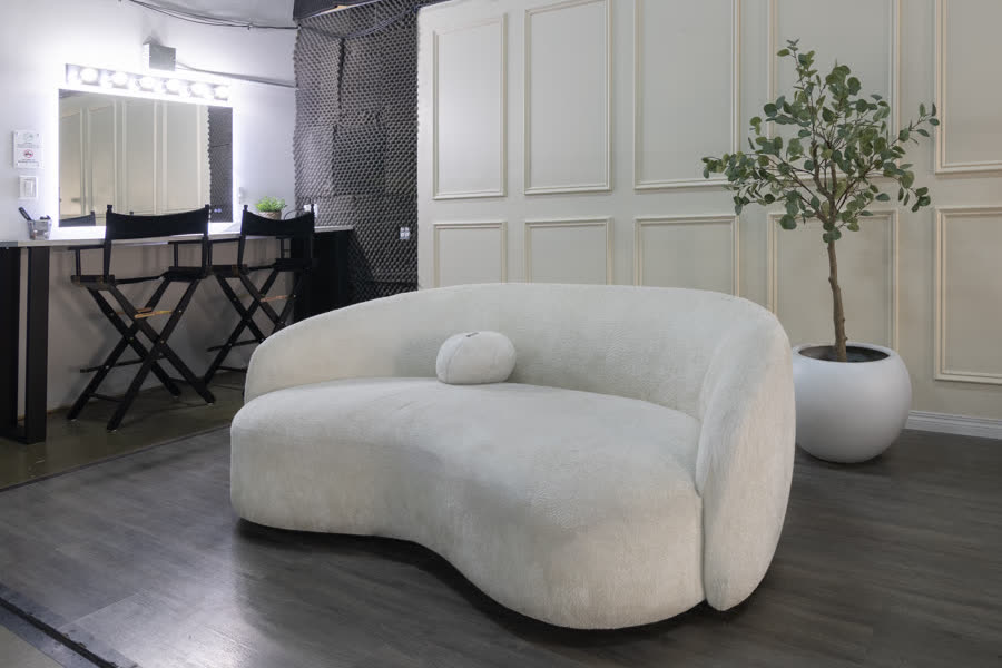 Studio A's living room set: a curved white boucle sofa and a lit vanity mirror against a panelled wall, with a potted tree