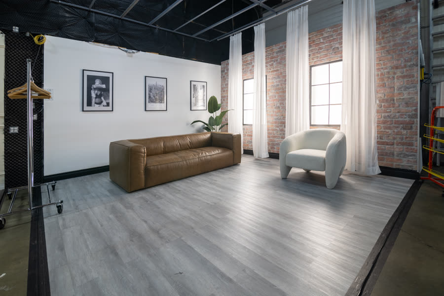 Studio A's New York apartment set: a brown leather sofa and a white boucle chair against exposed brick, framed prints and sheer curtains
